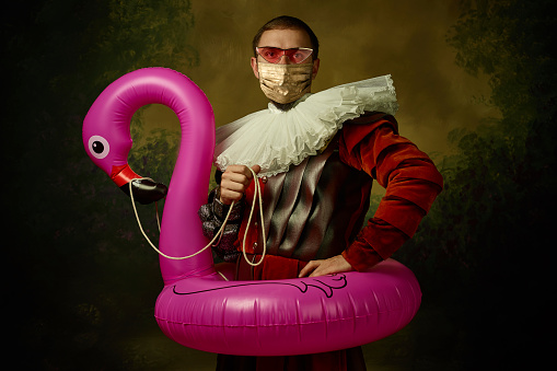 Portrait of medieval young man in vintage clothing, golden face mask and pink eyewear with flamingo on dark background. Royal person protected from covid. Concept of comparison of eras, modern, fashion.