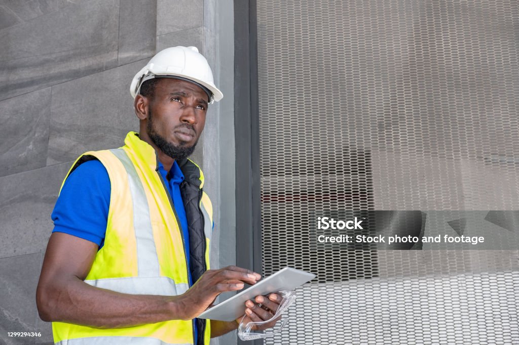 Selective focus at face of Black African foreman at building construction site, wearing protective hat and safety equipment while using digital tablet to record information. Civil engineer working. Building Contractor Stock Photo