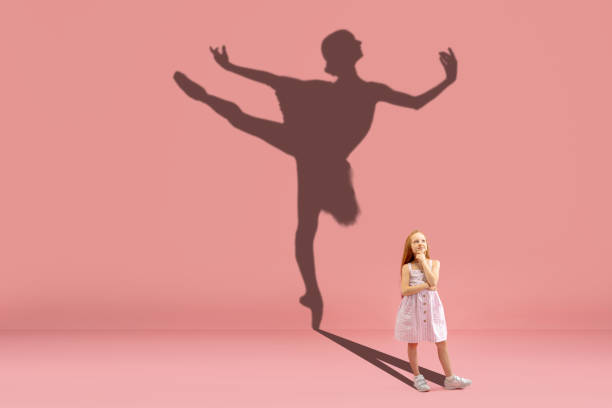 Childhood and dream about big and famous future. Conceptual image with girl and shadow of fit ballerina dancing on coral pink background Childhood and dream about big and famous future. Conceptual image with girl and drawned shadow of ballerina dancing on coral pink background. Childhood, dreams, imagination, education concept. ballerina shadow stock pictures, royalty-free photos & images