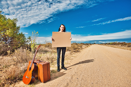 With her suitcase and guitar, a beautiful young woman waits for a lift at the roadside under an impressive summer sky.