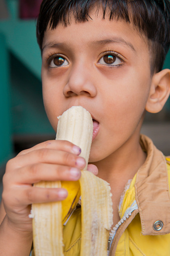 Indoor image of an Asian, Indian cute little boy in yellow clothes eating a fresh banana at home.