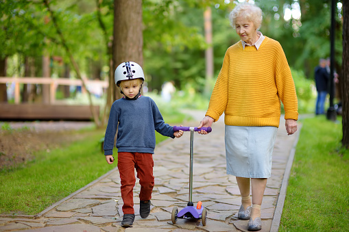 Beautiful granny and her little grandchild walking together in autumn park. Boy riding by scooter. Active family leisure.