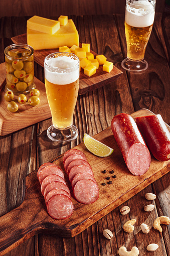 Sliced smoked salami on cutting board with two glasses of beer, cubs of cheese, olives, chestnuts and pistachios