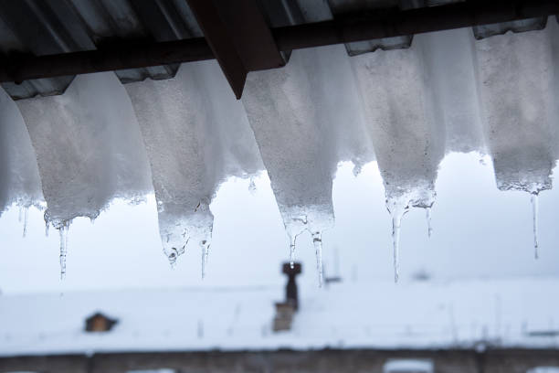 ghiaccio e neve coprono la costruzione del tetto, pericolose dighe di ghiaccio appese al tetto.  concetto di pericolo e sicurezza - icicle hanging snow moving down foto e immagini stock