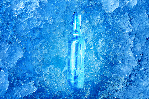 the vaccine in a glass ampoule with a clear liquid with a medical preparation for the treatment of a viral disease is cooled to subzero temperatures lying on frozen ice, blue background on pharmaceut