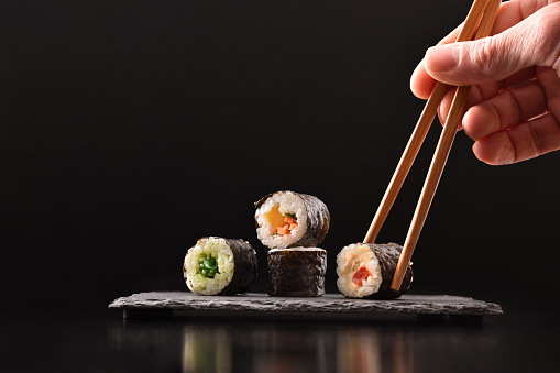 Background hands picking four assorted vegetable maki sushi with chopsticks on slate serving plate on dark wooden table with black background. Front view. Horizontal composition.