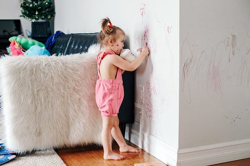 Divertido lindo bebé niña dibujo con marcador en la pared en casa. Niña niña pequeña con botella de leche jugando en casa. Auténtico momento de estilo de vida de la infancia. Joven artista pintando en la pared en la sala de estar photo