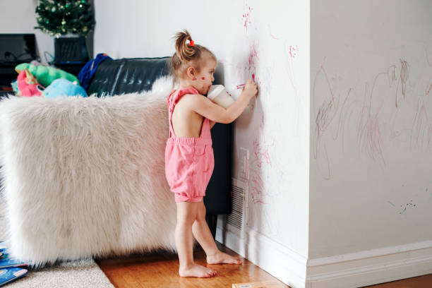 divertido lindo bebé niña dibujo con marcador en la pared en casa. niña niña pequeña con botella de leche jugando en casa. auténtico momento de estilo de vida de la infancia. joven artista pintando en la pared en la sala de estar - travesura fotografías e imágenes de stock