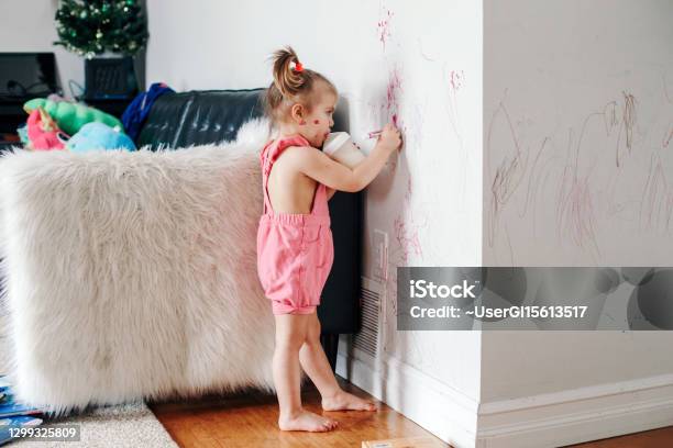 Funny Cute Baby Girl Drawing With Marker On Wall At Home Toddler Girl Child With Milk Bottle Playing At Home Authentic Candid Childhood Lifestyle Moment Young Artist Painting On Wall At Living Room Stock Photo - Download Image Now