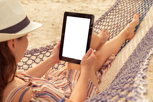 Woman relaxing while on vacation lying in a hammock on the beach, holding a tablet computer with blank screen