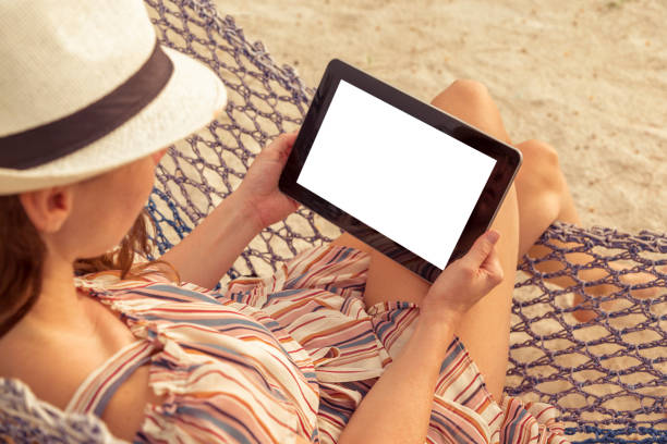 istock Woman holding a tablet computer while sitting in a hammock on the beach 1299330061