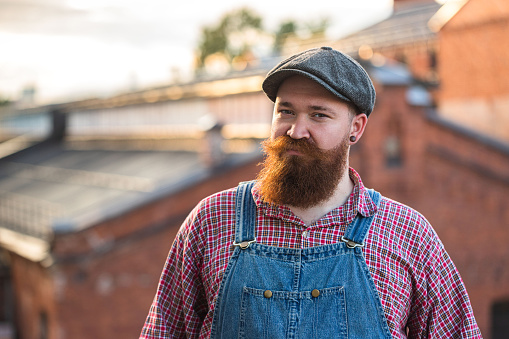 Portrait of brutal bearded stylish craftsman wearing blue overalls, checked shirt and cap in vintage style of the mid 20th century, looking at camera, outdoors. Family workshop, old manufactory worker