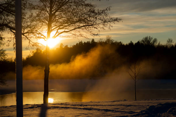 Horizontal composition photography of a large frozen lake in winter season, with a small white cabin in middle of forest with orange sunlight from twilight and some fog / mist due to humidity.