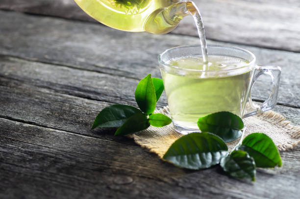 cette en verre de thé vert avec les lames fraîches de thé sur la table en bois, concept chaud de boisson - culture du thé photos et images de collection