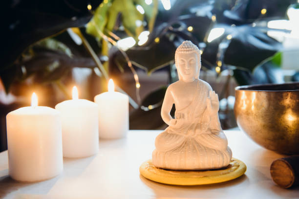 istock Decorative white Buddha statuette , burning candles and singing bowl with green monstera plant on the background. Meditation and relaxation ritual. Exotic massage. Wide banner. Selective focus. 1299349876