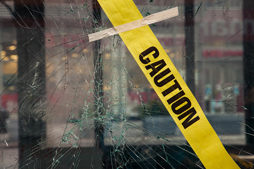 Cracks radiate from the center of the glass where the impact was made. Down town shop windows in the background in strong blur. Yellow warning tape with the text \