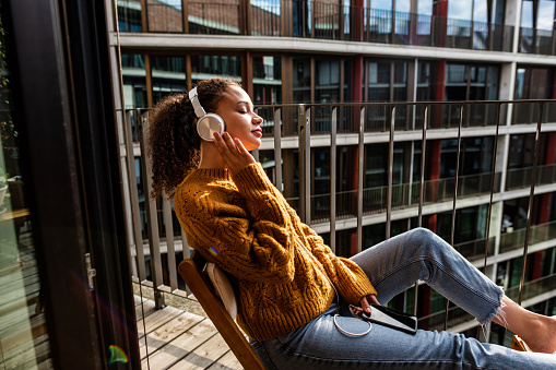 Side view of a Dominican young woman sitting on her balcony with headphones and enjoying a sunny day