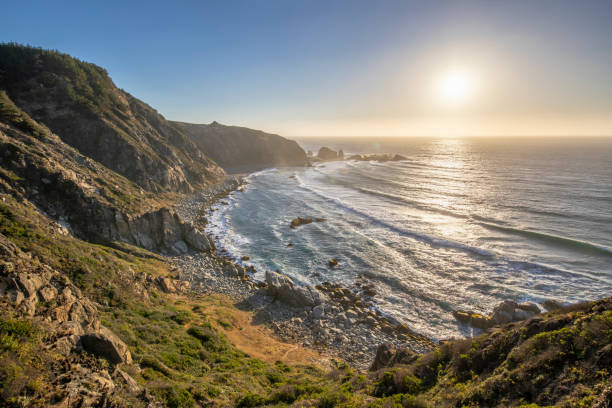 una puesta de sol asombrada en un paisaje de playa idílico con la puesta de sol sobre el agua y la luz del sol generando un ambiente de mal humor. un increíble paisaje de wil con los acantilados y las olas del mar salpicando agua - sky landscape horizon over water sunlight fotografías e imágenes de stock