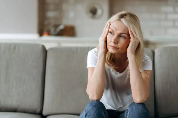 Photo of Sad caucasian senior woman wearing casual clothes sits on couch at home alone feels unhappy because of headache, stress, illness or bad news, she needs rest and sleep