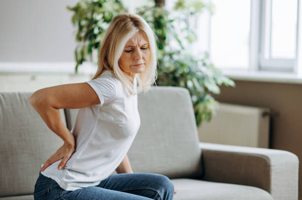 腰痛に苦しむ成熟した女性、リビングルームのソファに座っている不幸なシニアブロンド、背中の痛みのために不快感を感じる - arthritis osteoporosis pain backache ストックフォトと画像