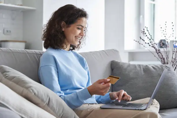 Photo of Happy hispanic young woman consumer holding credit card and laptop buying online at home. Female shopper customer shopping on ecommerce website market place making digital payment using bonus money.