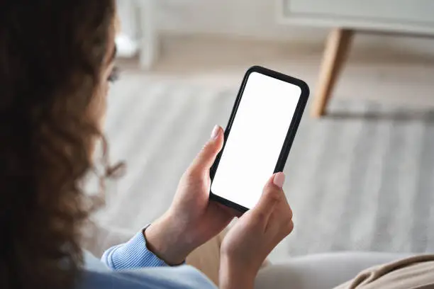 Photo of Young woman or teen girl hands holding smart phone with mockup white blank display, empty screen for social media app ads at home. Mobile applications technology concept, over shoulder close up view.