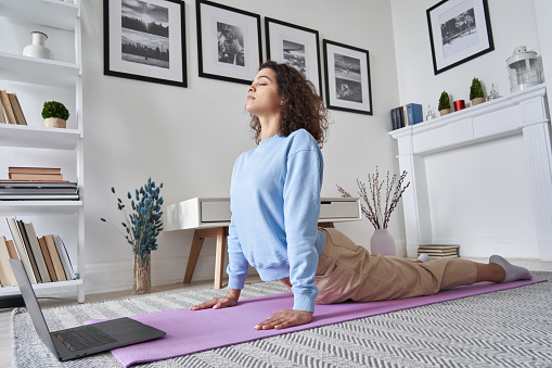 Fit young woman doing yoga exercise watching tv yoga class learning position with fitness tutorial on laptop computer stretching on mat at home, training body flexibility, online workout concept.