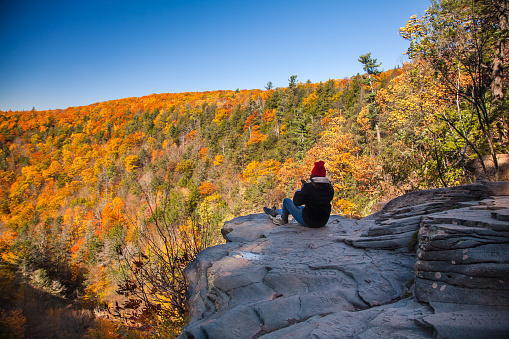 The person, tourist sits on the edge of a cliff and looks at the autumn, fall orange, red and yellow leaves, foliage, and trees in the mountains. Young woman in  jeans and jacket. Wearing a read hat.