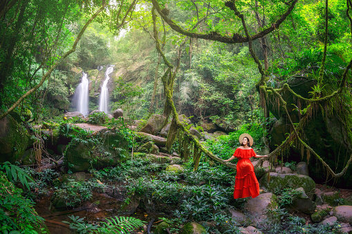 Beautiful woman in red dress sitting on the vine in front of the Sapan Waterfall, Khun Nan National Park, Boklua District, Nan Province, Thailand.