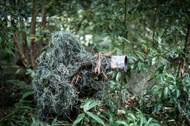 A photographer wearing a Ghillie suit is in a forest hiding and taking photos with a camera and telephoto lens.