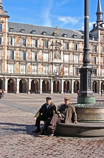 Two elderly Spanish men sitting on a bench with the facade of the Casa De La Panaderia to the rear, Plaza Mayor, Madrid, Spain.