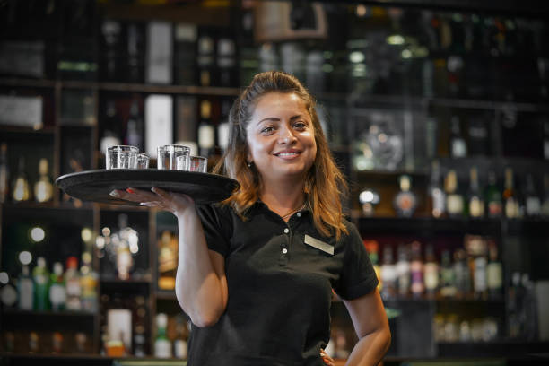 The waitress is carrying a whiskey glass. Waitress carries a whiskey glass on a tray in hotel restaurant, bar. The concept of service. Shelves with bottles of alcohol in the background. Night time. waitress or barmaid stock pictures, royalty-free photos & images