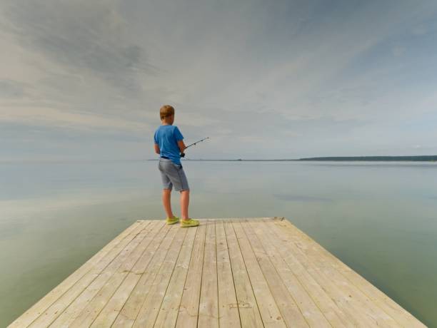 piccolo ragazzo dai capelli biondi sta pescando sulla talpa. bambino in camicia blu, pantaloncini a righe grigie e infradito verdi - sweden fishing child little boys foto e immagini stock