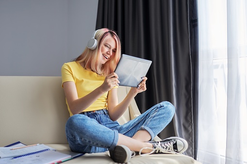 Teenager girl student in headphones looking in screen of digital tablet, sitting at home on couch. Video chat, online lesson, e-learning, female talking and learning remotely