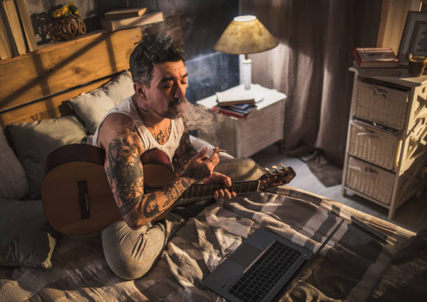istock hipster man with tattoo sitting on bed and playing acoustic guitar, 1299403469