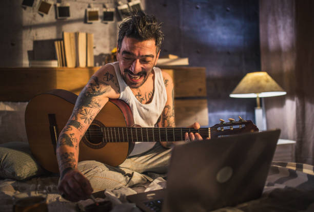 istock hipster man with tattoo sitting on bed and playing acoustic guitar, 1299403520