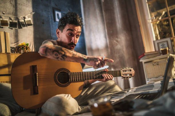 istock hipster man with tattoo sitting on bed and playing acoustic guitar, 1299403588