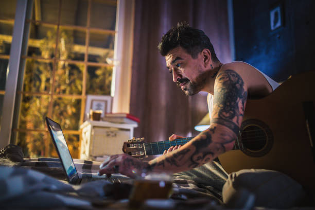 istock hipster man with tattoo sitting on bed and playing acoustic guitar, 1299403633