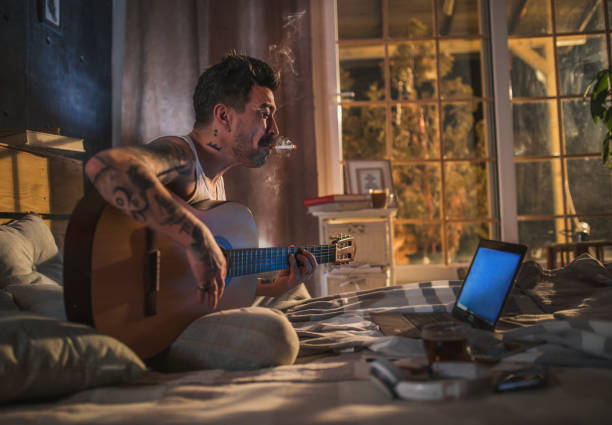 istock hipster man with tattoo sitting on bed and playing acoustic guitar, 1299403707