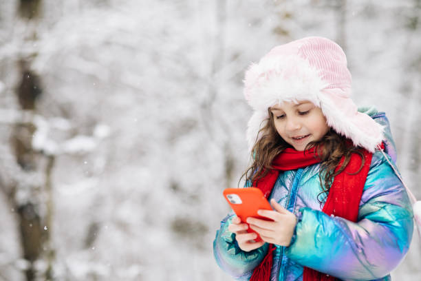 istock Happy little kid use smartphone funny face mask app look at cell screen smiles at something funny. Holidays and celebrations concept. Outside. The dead of winter, cold weather, frost, snow 1299404691