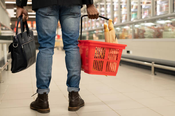 Rear view of low section of young man with red basket moving between displays Rear view of low section of young man with red basket moving between large and long displays with frozen food in modern supermarket holding shopping basket stock pictures, royalty-free photos & images