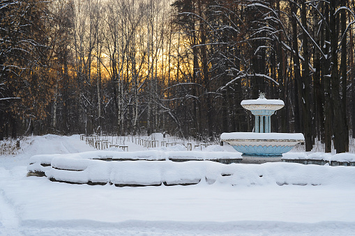Old abandoned park with fountain. Winter landscape with forest background and sunset.