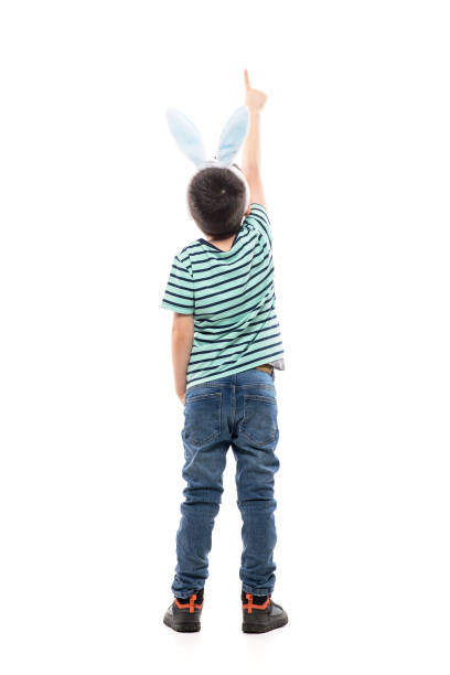 Behind view of young boy with Easter bunny ears showing direction pointing finger up. Full length isolated on white background.
