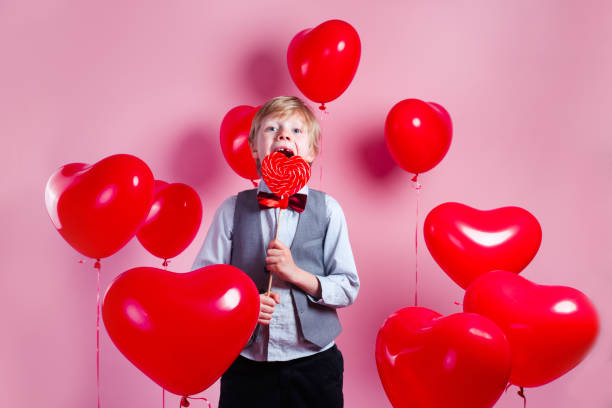 バレンタインデー。赤いハート風船の背景にキャンディーを食べる小さなかわいい男の子 - child valentines day candy eating ストックフォトと画像