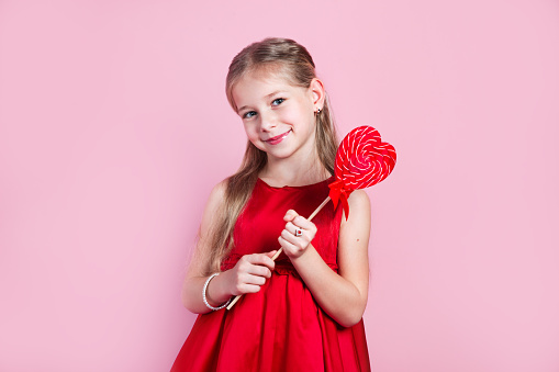 Valentines day. Little cute girl in red dress with a heart-shaped lollipop on pink background