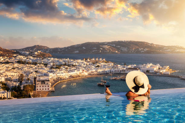 A woman with a glas of wine in a swimming pool enjoys the view over the town of Mykonos island, Greece A tourist woman with a glas of wine in a swimming pool enjoys the view over the town of Mykonos island, Greece, during summer sunset time greek culture photos stock pictures, royalty-free photos & images