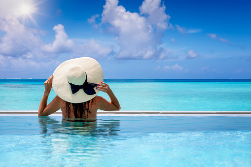 A woman with a big sunhat enjoys the view from the swimming pool to the turquoise sea of the Indian Ocean during her tropical holidays