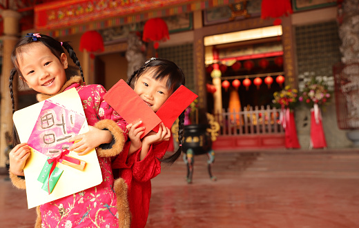 two asian twins girls wearing cheongsam make a New Year’s visit. They holding New Year’s Money and Spring couplets. Traditional custom of chinese new year.