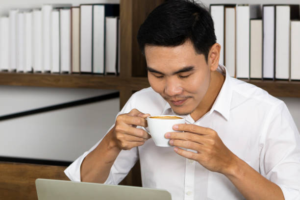 istock Young man smelling aroma of coffee in the cafe before drinking. Man drinking coffee with coffee cup in morning time. Handsome young man smelling his coffee sitting in his room. 1299421328