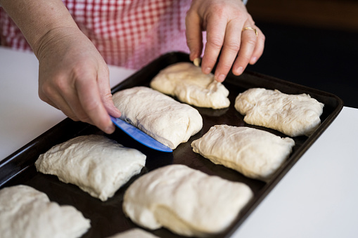 Close view of Caucasian woman using scraper to set up individual pieces of dough on baking sheet for ciabatta rolls.